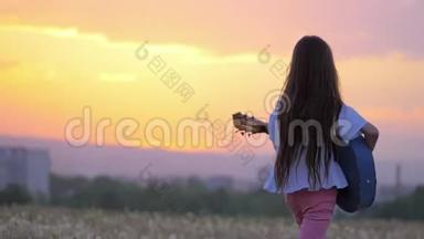 日落时美丽的风景。 有趣的小女孩音乐家弹吉他走在户外蒲公英田野远离城镇
